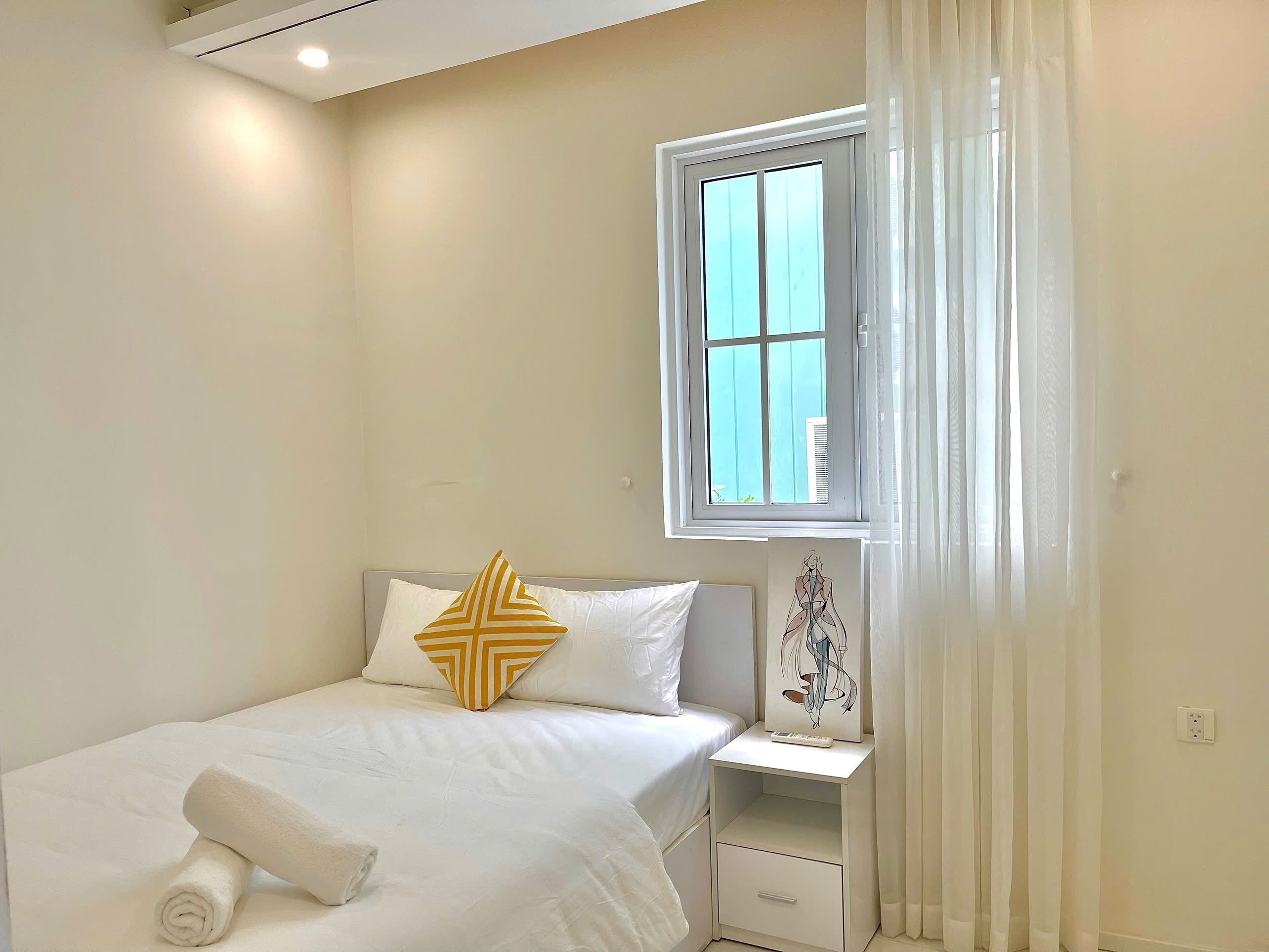 Una cama bien hecha con ropa blanca y una toalla enrollada, una mesita de noche con lámpara, y una ventana con cortinas.