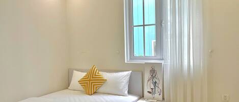 Una cama bien hecha con ropa blanca y una toalla enrollada, una mesita de noche con lámpara, y una ventana con cortinas.