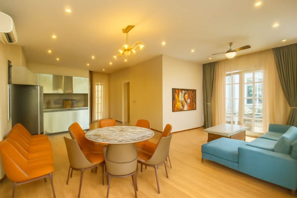 A modern living room with a round table, orange chairs, a blue sofa, and a kitchen area with a refrigerator and cabinets.