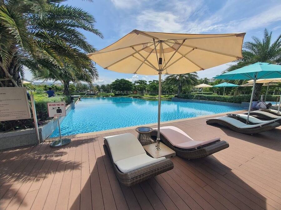 Un área al lado de la piscina con sillones de descanso y sombrillas, una piscina azul cristalina y palmeras.