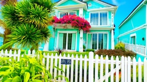 Una casa a due piani con un recinto bianco a stecche, una facciata color turchese e un portico adornato da piante fiorite rosa.