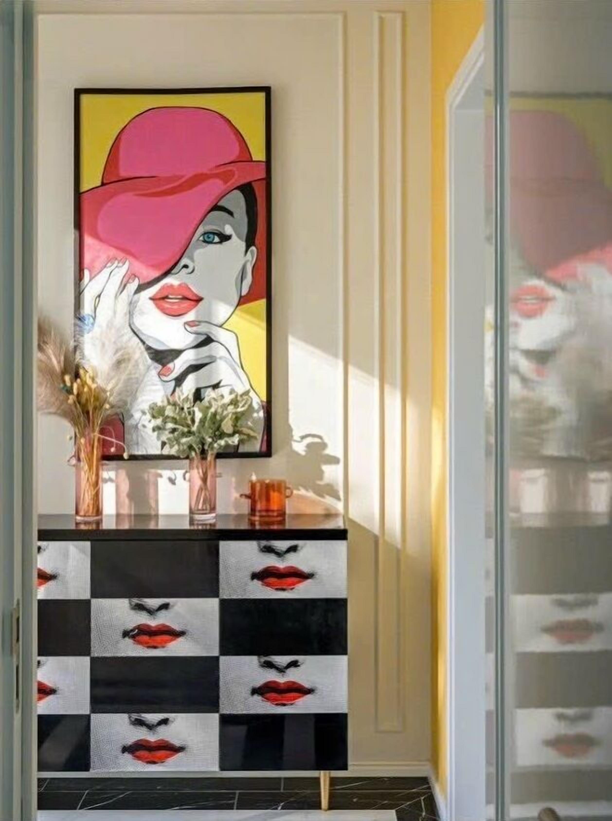A framed pop art painting on a wall, a checkered cabinet with red lips design, and a vase with dried flowers.