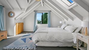 Ein Schlafzimmer mit schrägen Wänden, einem Bett, einem Nachttisch mit Lampe, einem Fenster mit Vorhängen und einem kleinen runden Spiegel an der Wand.