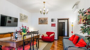 Ein Essbereich mit Holztisch, Stühlen und einer Blumenvase. Ein rotes Sofa und ein an der Wand befestigter Fernseher sind ebenfalls vorhanden.