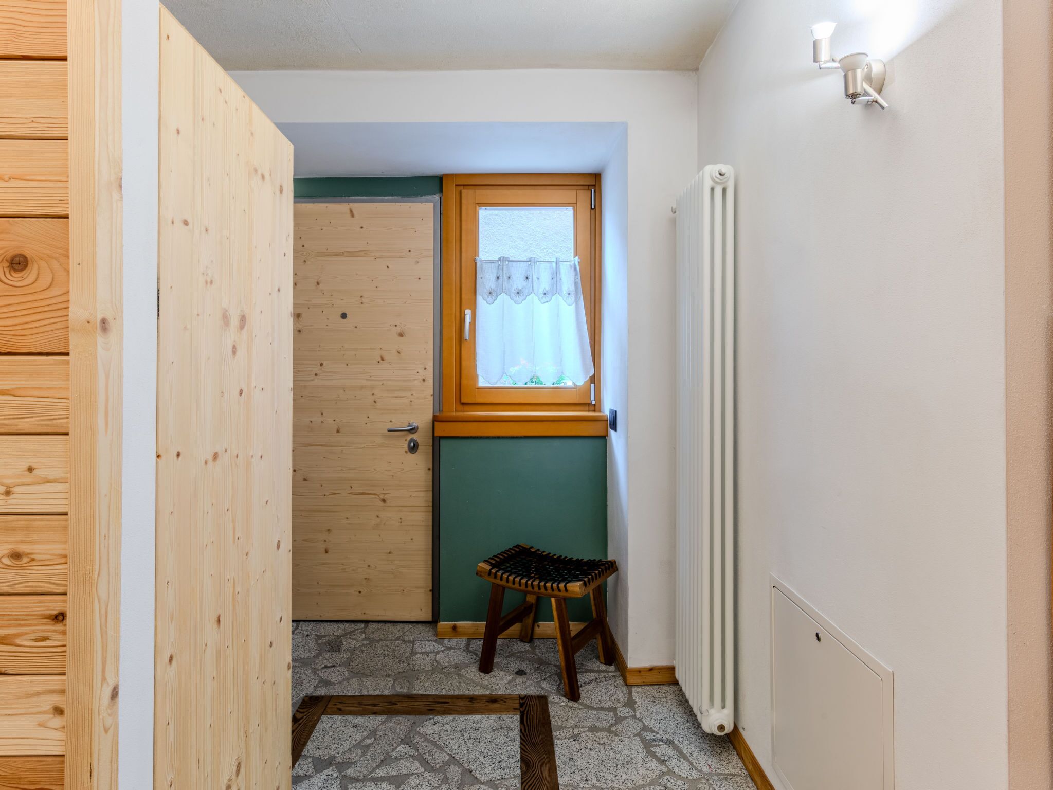 A small room with a wooden door, a window with a curtain, a radiator, and a small stool.