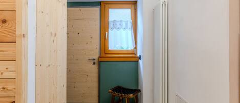A small room with a wooden door, a window with a curtain, a radiator, and a small stool.