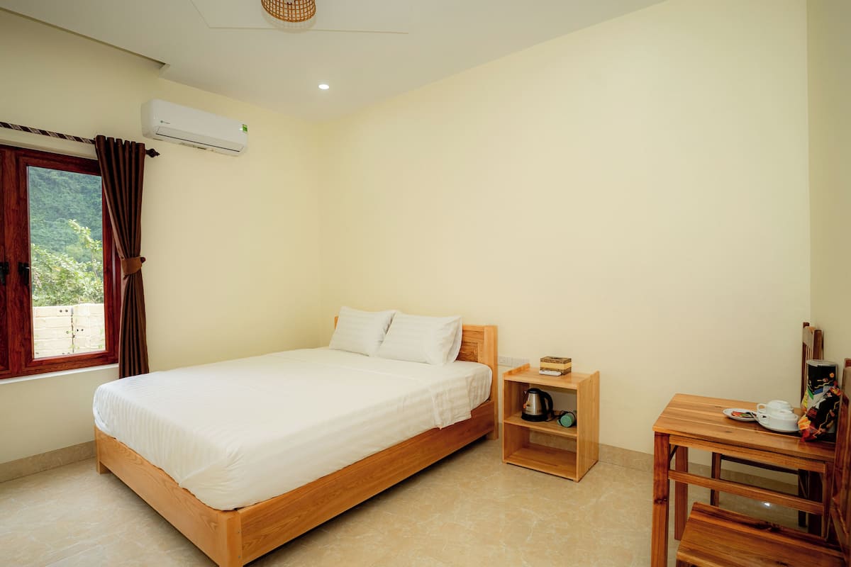 A bedroom with a wooden bed, a small wooden table with a kettle, and a window with curtains.