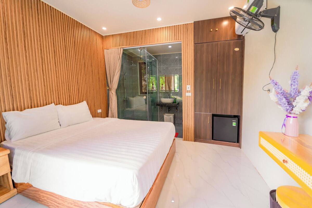 A modern bedroom with a large bed, a glass shower enclosure, wooden paneling, and a small TV.