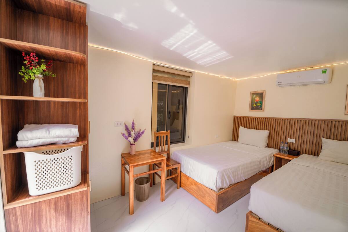 A hotel room with a wooden desk, two beds, a window, and an air conditioning unit.