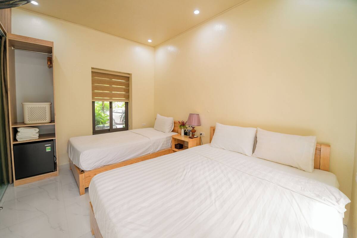 A hotel room with two beds, a wooden wardrobe, a small table with a lamp, and a window with blinds.