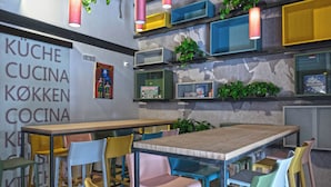 Un coin repas avec une table en bois et des chaises colorées, des étagères avec des plantes et des jeux de société, et un mur sur lequel est affiché un panneau indiquant : « KÜCHE KÖKKEN COCINA KÖKEN ».