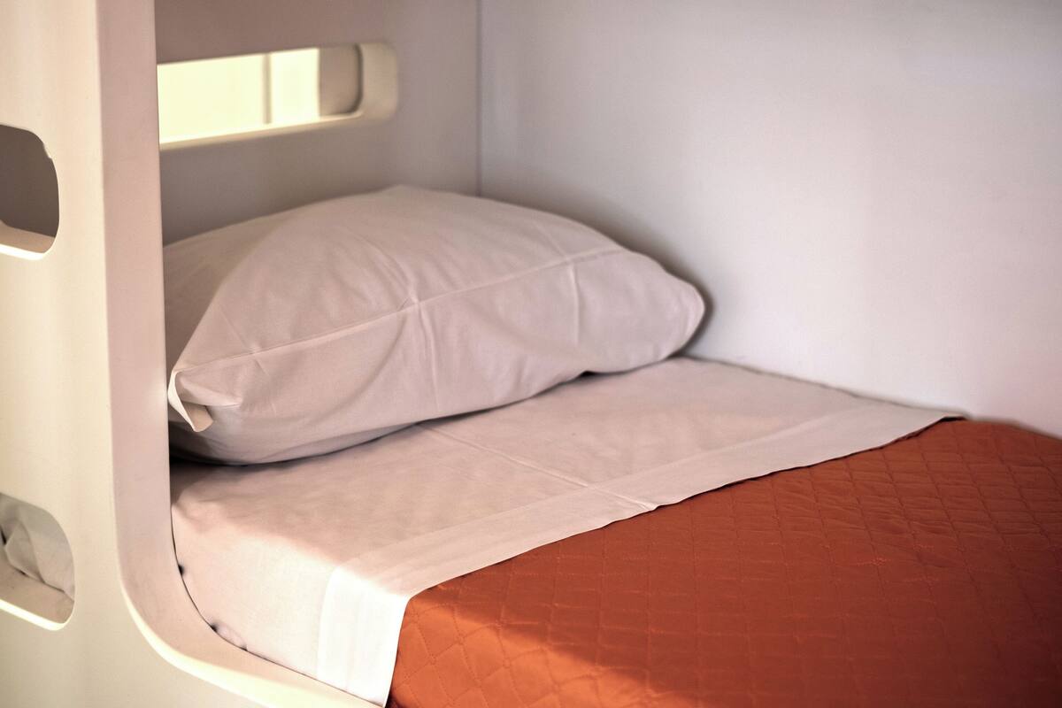 Quarto pequeno com uma cama de solteiro, cabeceira branca, colchão marrom e travesseiro branco.