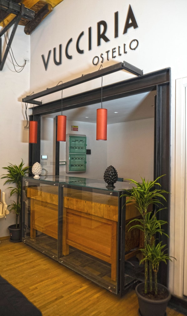 A reception desk with a glass top, wooden panel, and two red pendant lights. There are green electrical boxes and a potted plant on the desk.