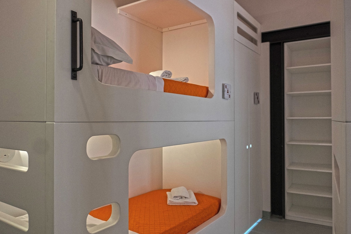 Uma cama beliche com cabeceira branca e roupa de cama laranja, uma parede branca com maçaneta, e uma porta branca com moldura preta.