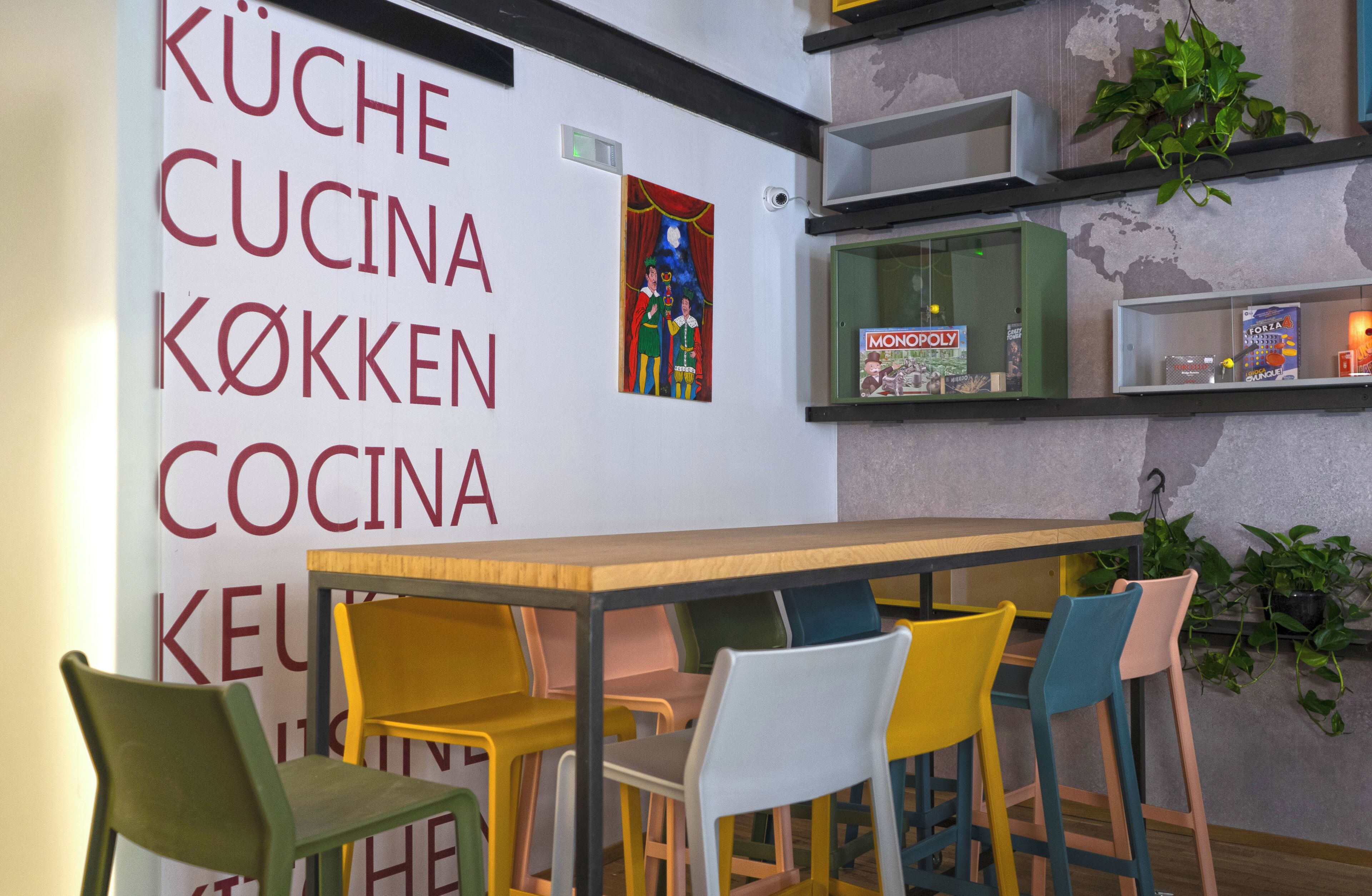 Un comedor con una mesa de madera y sillas coloridas, una pared con un letrero en varios idiomas, y estantes con libros y plantas.