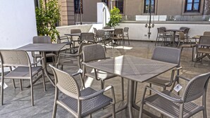 Un espace extérieur aménagé en terrasse, avec des tables et des chaises en métal, un pot de fleurs et un bâtiment en arrière-plan.