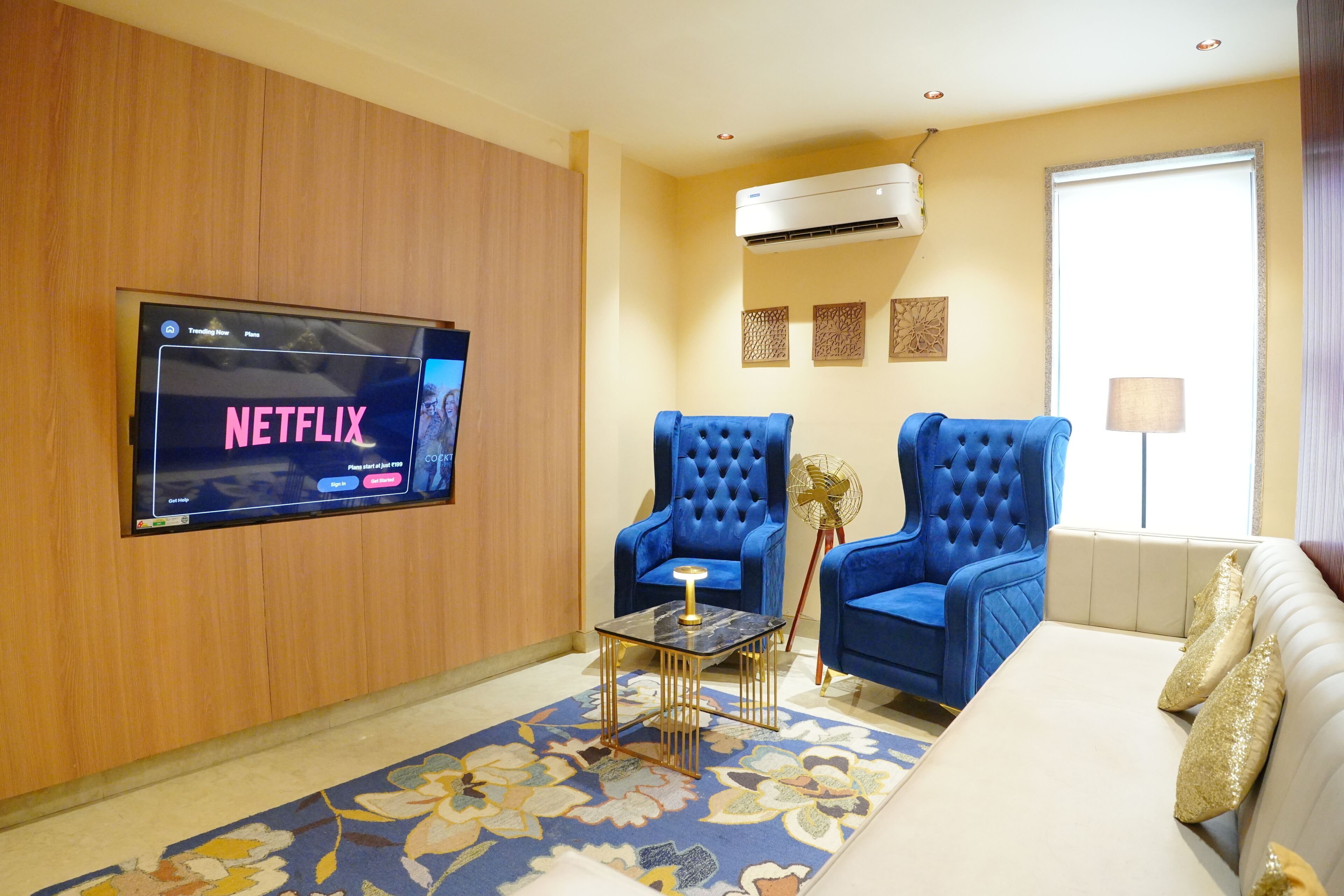A living room with a flat-screen TV mounted on the wall, two blue armchairs, a beige sofa, a small table with a lamp, and a decorative fan.