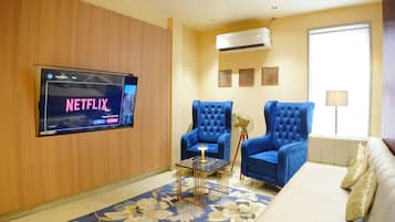 A living room with a flat-screen TV mounted on the wall, two blue armchairs, a beige sofa, a small table with a lamp, and a decorative fan.