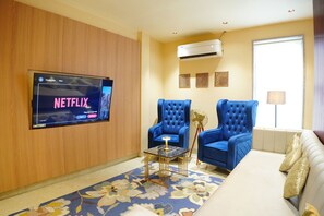 Una sala de estar con una televisión de pantalla plana montada en la pared, dos sillones azules, un sofá color beige, una mesita con una lámpara y un abanico decorativo.