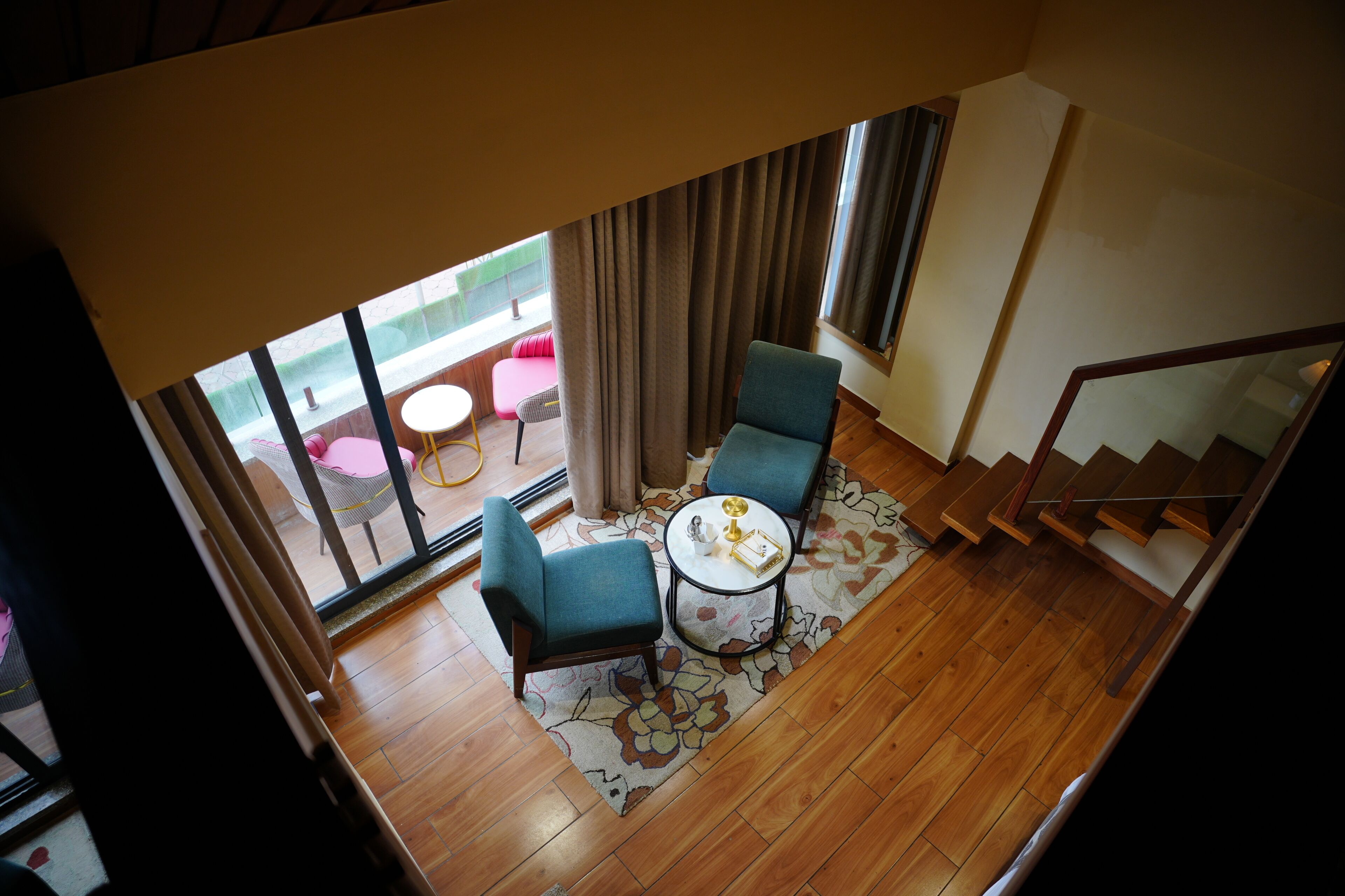 A room with a wooden floor, a round table with chairs, a view through a window with curtains, and a staircase.