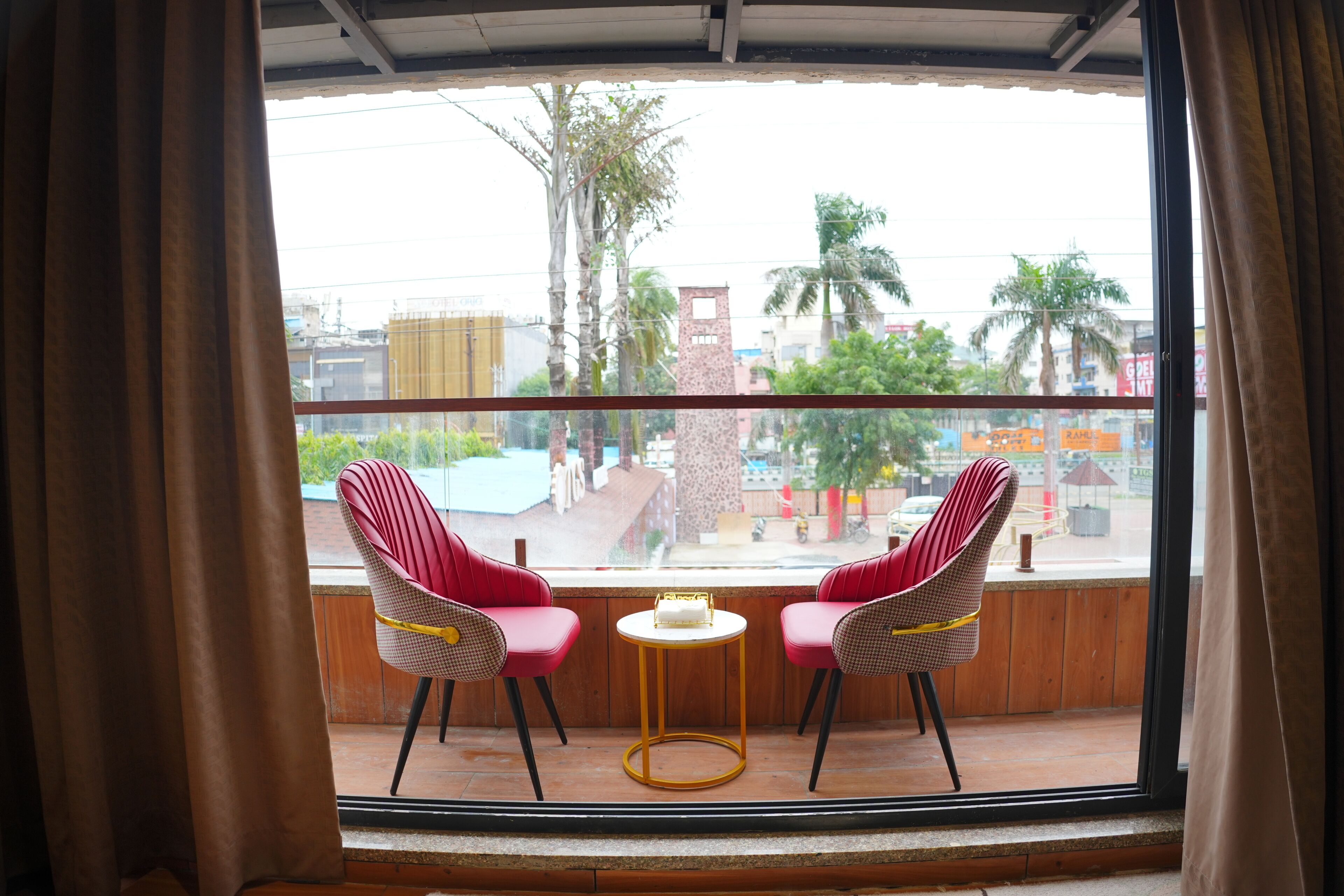 A balcony with two red chairs and a small round table, overlooking a street with buildings and palm trees.