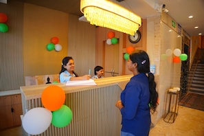 A hotel reception desk with a clock, a bulletin board, and a staircase in the background. - The Grand Sun City Hotel & Resort (Bhopal)