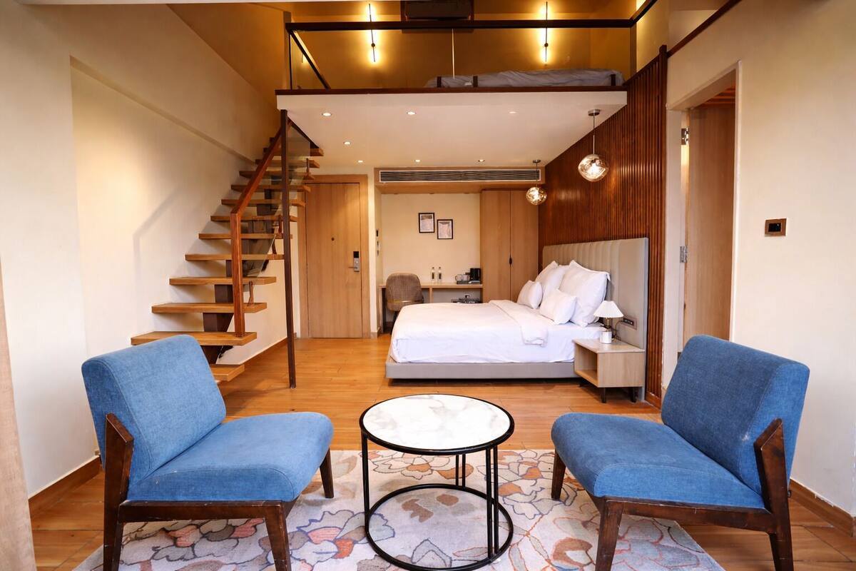 A bedroom with a bed, two blue chairs, a small round table, a staircase, and a wooden ceiling.