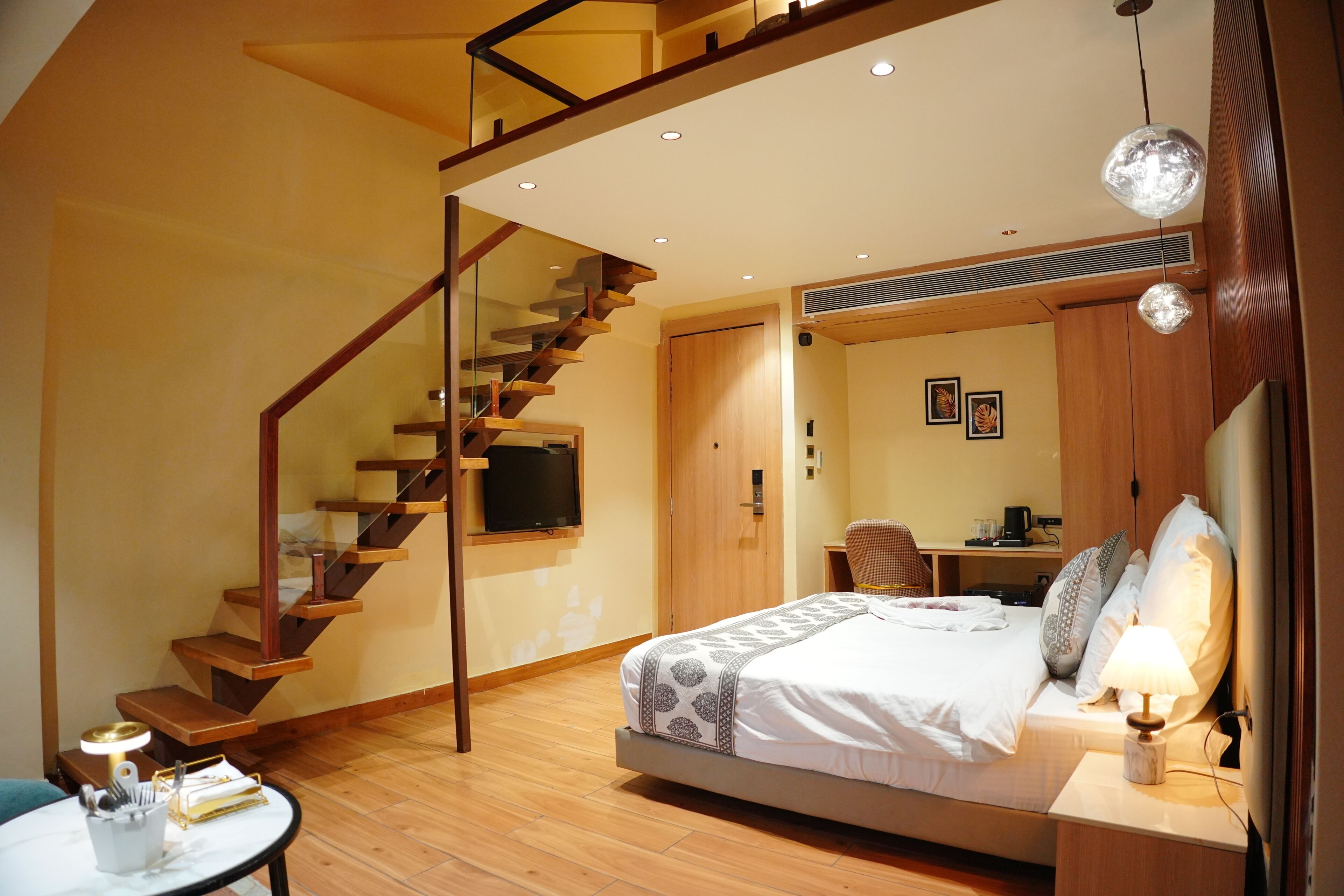 A modern hotel room with a wooden staircase, a bed with patterned bedding, a desk with a chair, and a television.