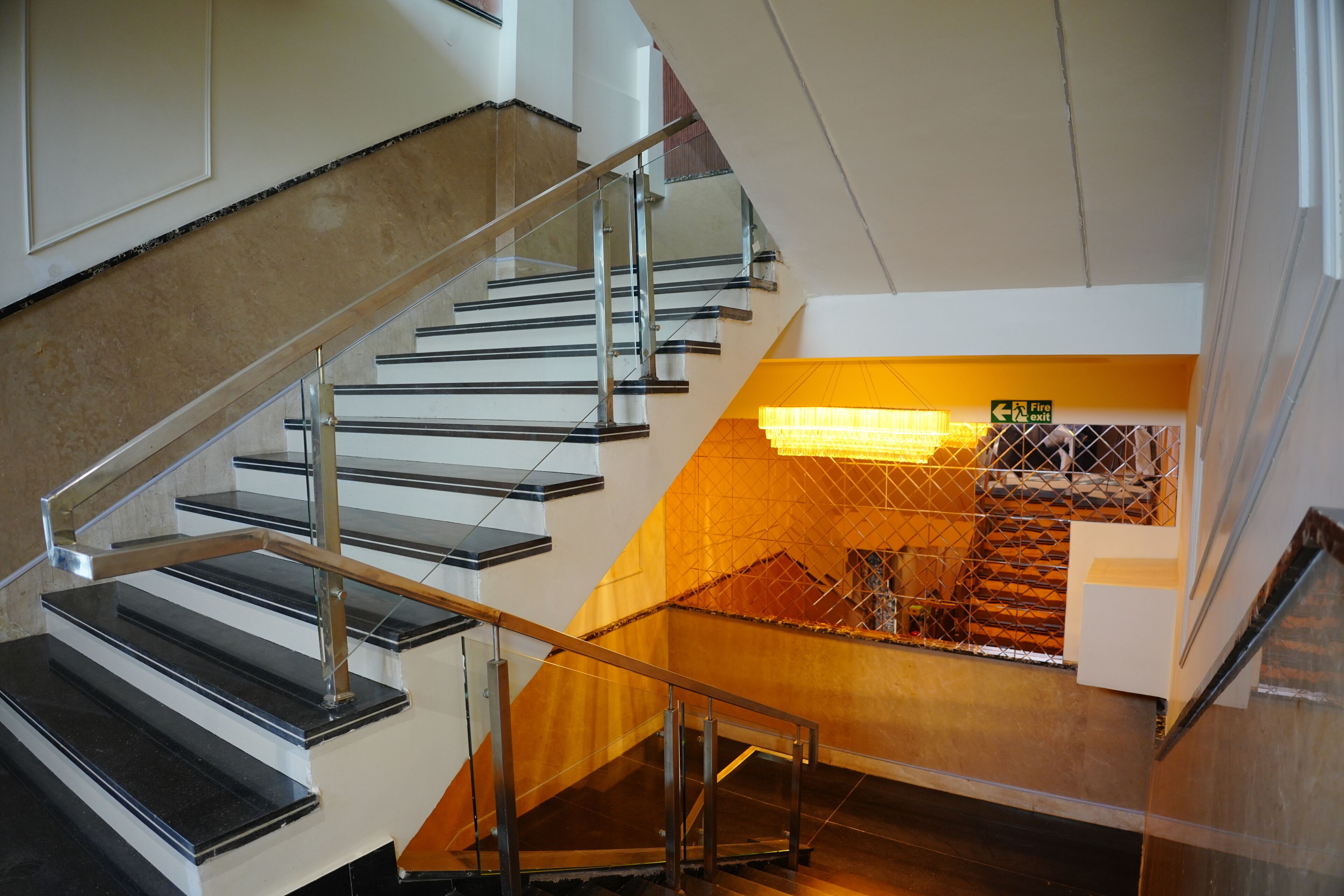 A staircase with metal railings and a glass barrier, leading to a lower level with a yellow-lit area and a fire exit sign.