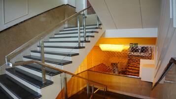A staircase with metal railings and a glass barrier, leading to a lower level with a yellow-lit area and a fire exit sign.