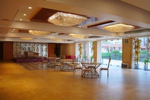A spacious hall with multiple seating arrangements, including round tables and chairs, and a stage area with a sofa and decorative backdrop.