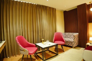 A hotel room with a large window, two red chairs, a small table, a sofa, and a lamp. - The Grand Sun City Hotel & Resort (Bhopal)