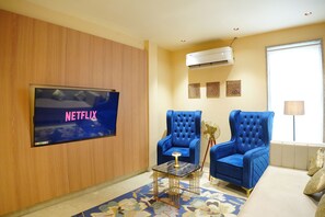 A living room with a flat-screen TV mounted on the wall, two blue armchairs, a small coffee table, and a patterned rug. - The Grand Sun City Hotel & Resort (Bhopal)