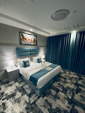 Une chambre d’hôtel moderne avec un grand lit, une tête de lit, une table de chevet avec une lampe, un tableau au mur et une moquette à motifs.