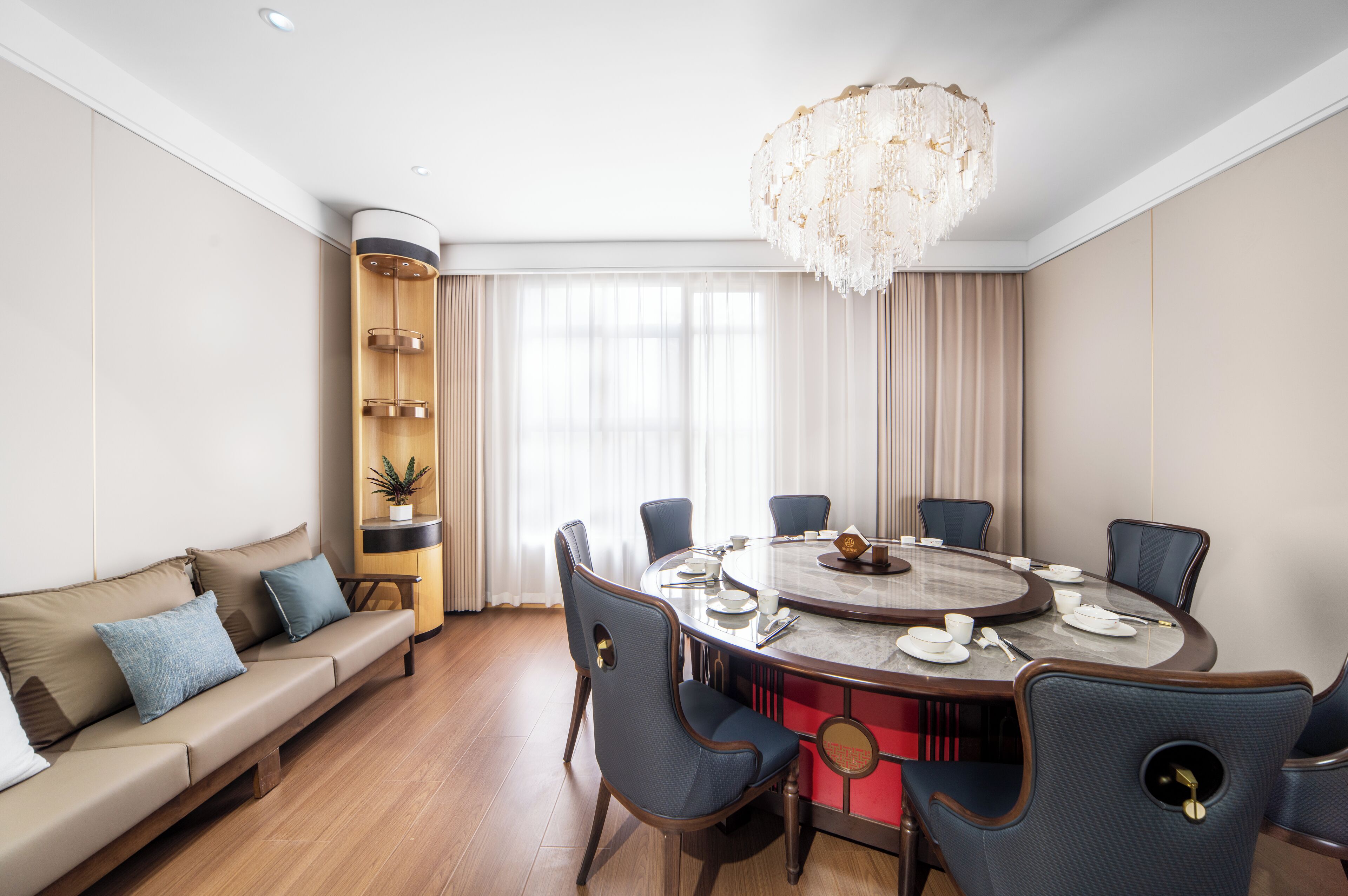 A modern dining room with a round table, six chairs, a chandelier, a sofa with cushions, and a bookshelf.