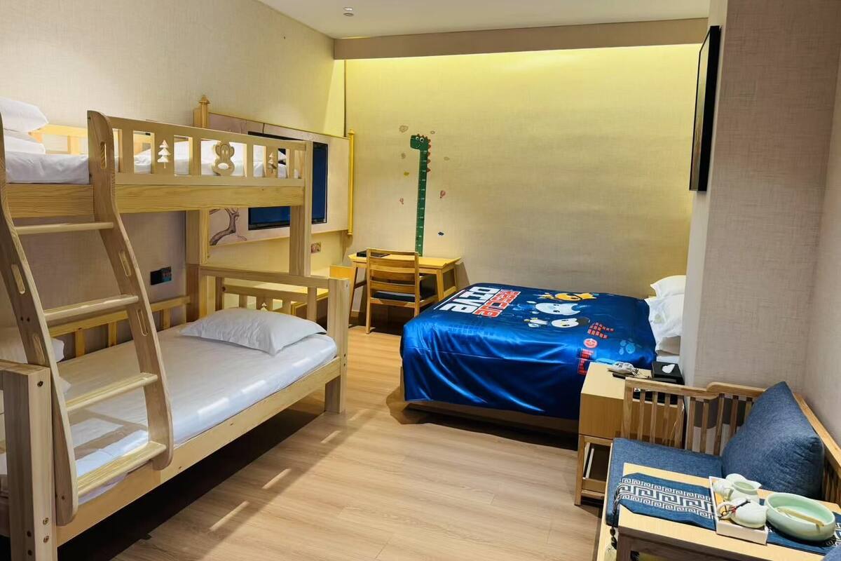 Egy kis méretű szállodai szoba emeletes ággyal, íróasztallal, széssel, egy kis asztallal, melyen egy lámpa található, és egy ággyal, melyen kék takaró van.