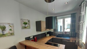 Eine kleine Küche mit Holztischplatte, Spüle und einem Stuhl. An der Wand befinden sich zwei Blumentöpfe, ein schwarzes Pendelleuchten und ein Fenster mit Vorhängen.