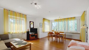 Een woonkamer met een bank, een salontafel, een tv-meubel, een eettafel met stoelen en grote ramen met gordijnen.
