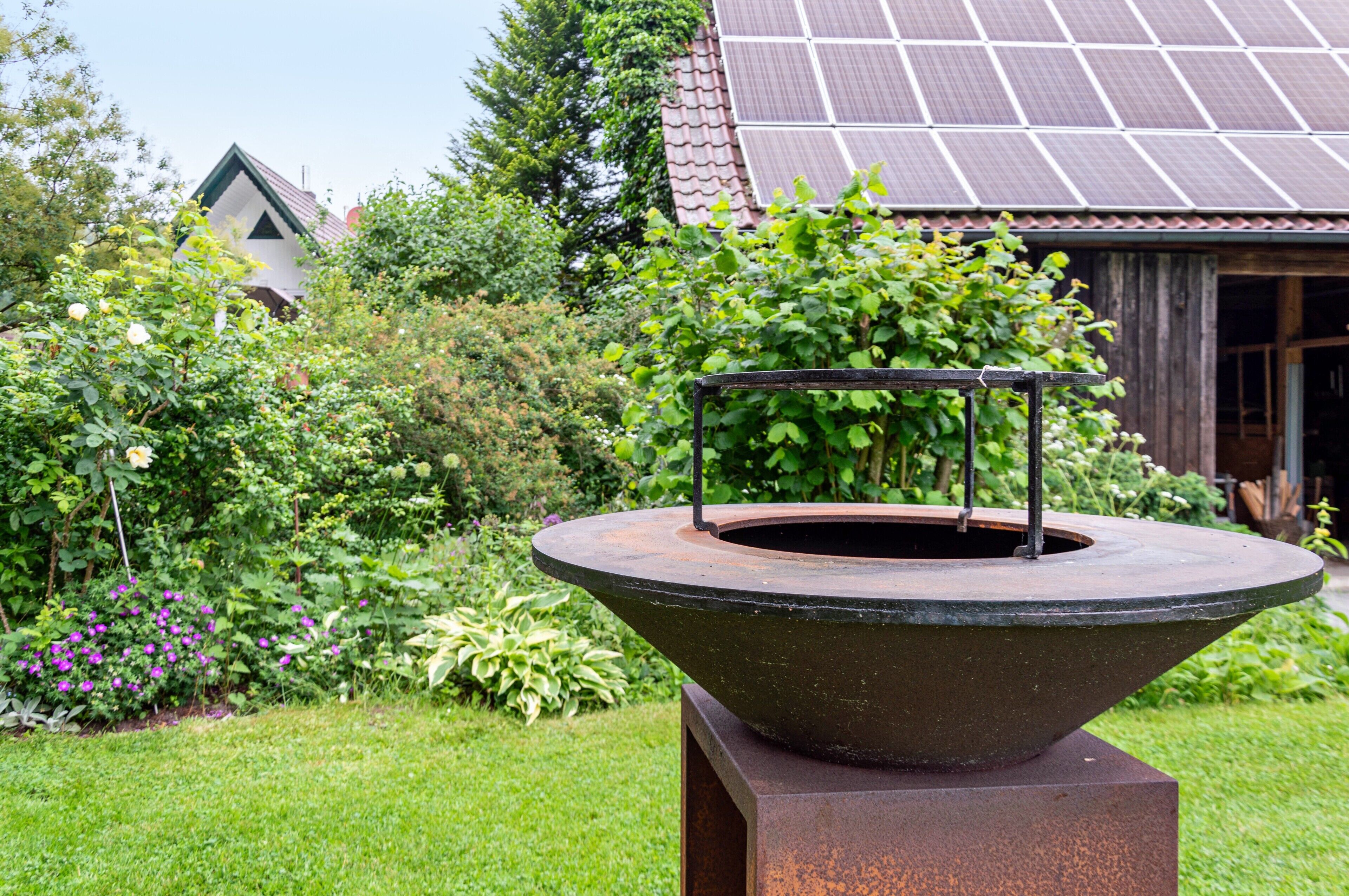 Ein Garten mit einem großen Metallpflanzgefäß, ein Gebäude mit einer Solardach-Anlage und üppigem Grün.