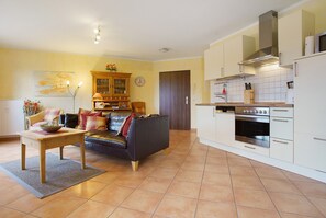 A living room with a sofa, a wooden table, and a kitchen with white cabinets and an oven.