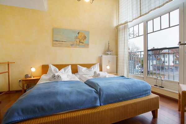 A bed with a blue blanket, a painting on the wall, and a window with a view of buildings.