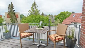 Un balcon avec une table et deux fauteuils en osier, des plantes en pot et une vue sur les bâtiments et les arbres.