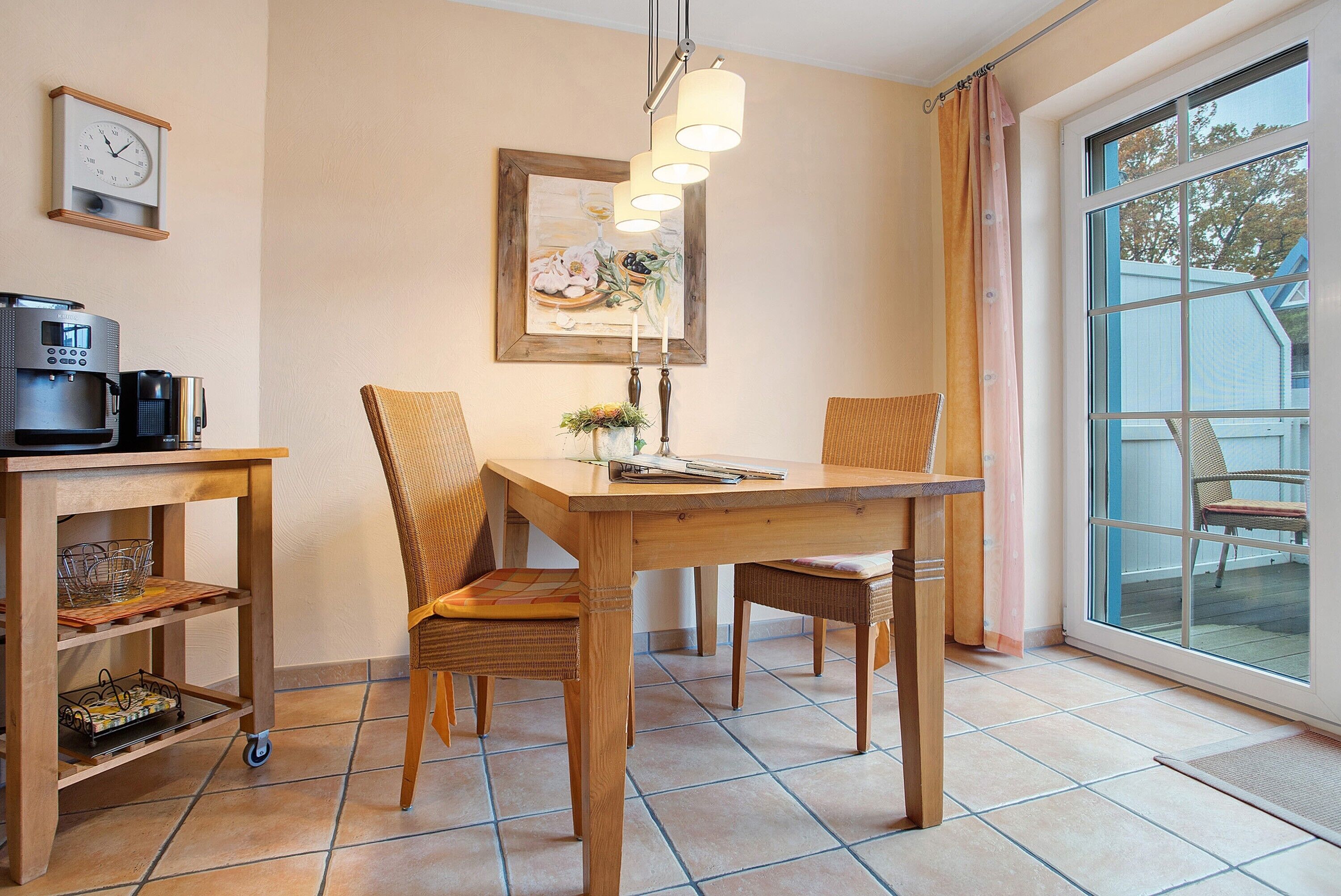 Un coin repas comprenant une table en bois, des chaises, une machine à café, une œuvre d’art encadrée et une fenêtre avec des rideaux.