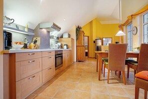 A kitchen with wooden cabinets, a stove, and a dining area with wicker chairs and a table. - Wellness & Living Under Thatch: Apartment 18 'Wellness Residence' (Zingst)