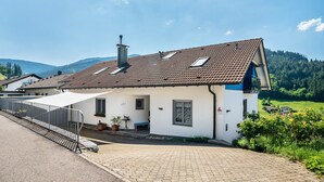 Ein Haus mit einem Ziegeldach, einem Schornstein, einer gepflasterten Auffahrt und einer kleinen Veranda mit einer Bank.