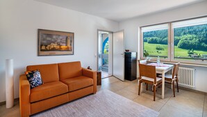 Ein modernes Wohnzimmer mit einer Couch, einem Esstisch mit Stühlen, einem Heizkörper und einem großen Fenster mit Blick auf Grünflächen.