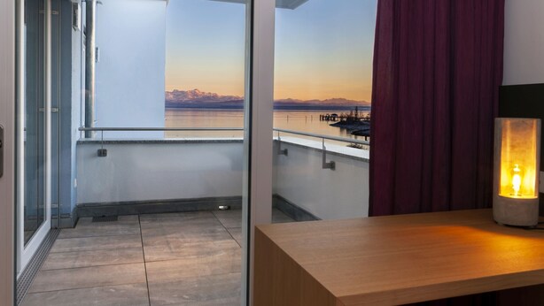 A balcony with a view of a body of water and mountains, a wooden table with a light, and a red curtain. - Apartment 'South-West Lake View' – Exclusive Lake Constance Retreat (Immenstaad am Bodensee)