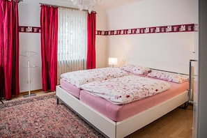 Un dormitorio con cama, cortinas, lámpara, alfombra y un adorno en la pared.