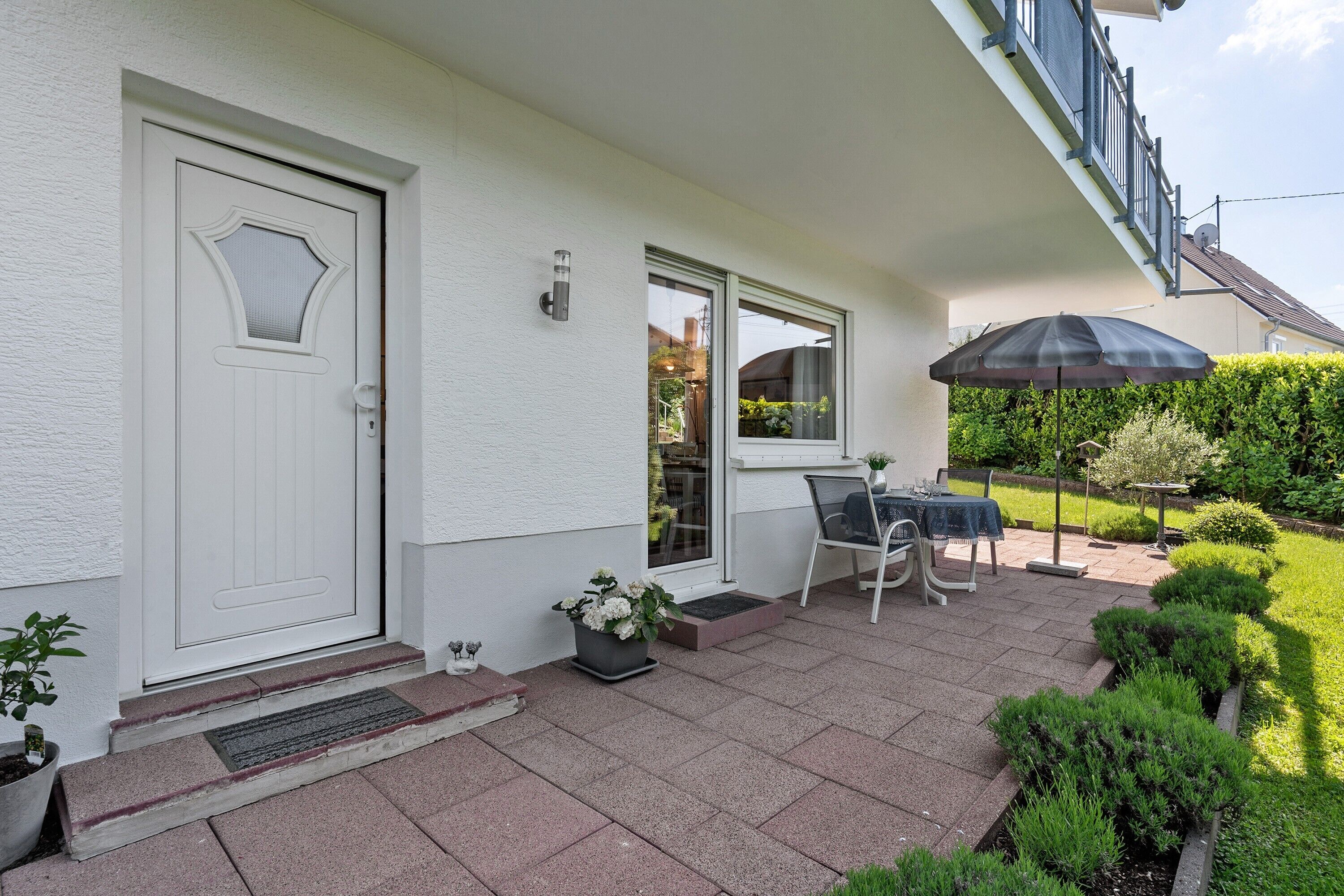 A house with a white door, a small porch with a table and chairs, and a garden with trimmed bushes.