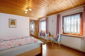 Ein Zimmer mit Holzboden, einem Bett mit floralem Bettzeug, einem kleinen Tisch mit Stuhl und einem Fenster mit Vorhängen.